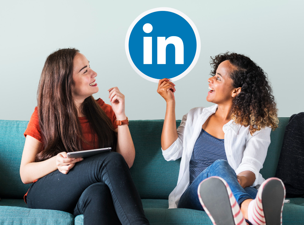 Two women on a couch, both looking at the LinkedIn logo one woman is holding